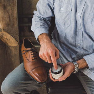 Man is putting his fingertips into a jar of Pure Polish Leather Cleaner and Conditioner, as he's about to condition his light brown leather oxfords