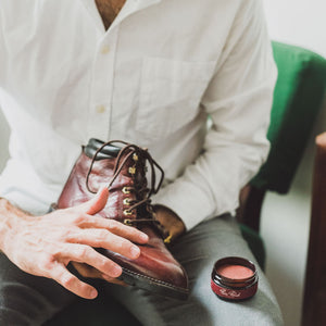 Man applies burgundy cream polish to the toe of a reddish lace up leather boot with his fingertips