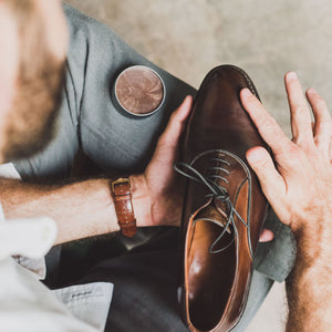 Man polishes the toe of a single brown leather oxford shoe, using an open tin of brown wax polish